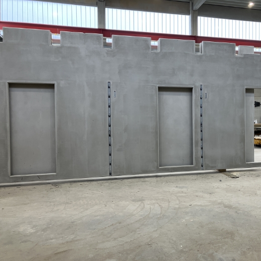 Großes, graues Carbonbeton-Fertigteil für eine Wand, das zwei Aussparungen für Türen sowie eine zinnenartige Oberkante aufweist, in einer Fertigungshalle stehend.