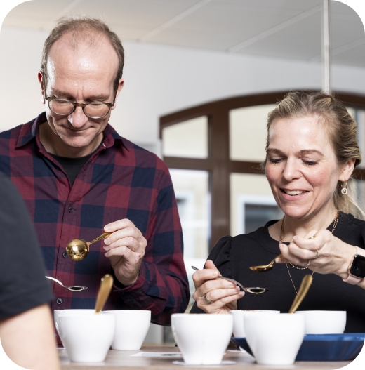 Nahaufnahme eines Mannes mit Brille und rot-kariertem Hemd und einer Frau, die goldene Löffel in kleine, weiße Cupping-Schalen halten, um Kaffee zu verkosten (Cupping).