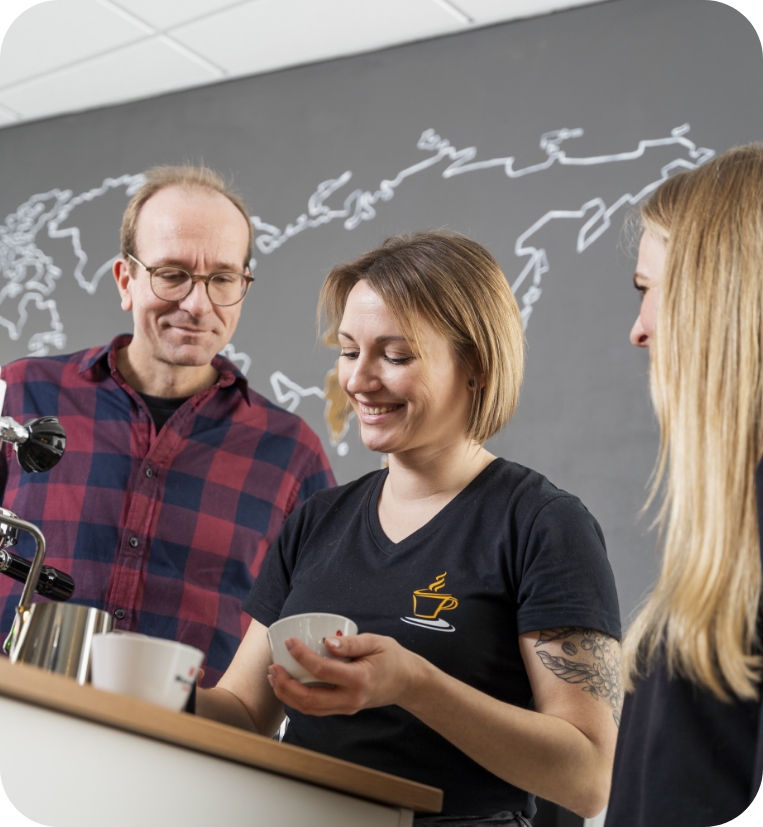 Drei Mitarbeiter von JoKa Kaffee, eine Frau mit tätowiertem Arm in einem schwarzen Logo-Shirt hält eine Tasse und lächelt. Links steht ein Mann mit Brille und rot-kariertem Hemd. Im Hintergrund ist eine Weltkarte als Kreidezeichnung an der dunkelgrauen Wand zu sehen.