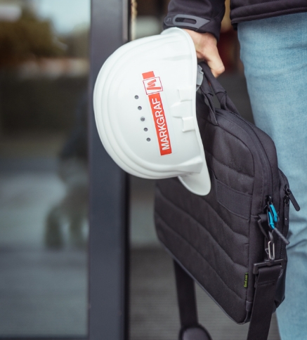 Nahaufnahme eines weißen Schutzhelms mit rotem Markgraf-Logo, der zusammen mit einer schwarzen Laptoptasche von einer Person gehalten wird, die eine Jeans trägt.