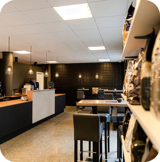 Innenansicht des JoKa Kaffee Ladengeschäfts mit einer dunklen Theke, einem Sitzbereich mit Stehtischen und Barhockern sowie einem Regal voller Kaffeeprodukte im rechten Vordergrund.