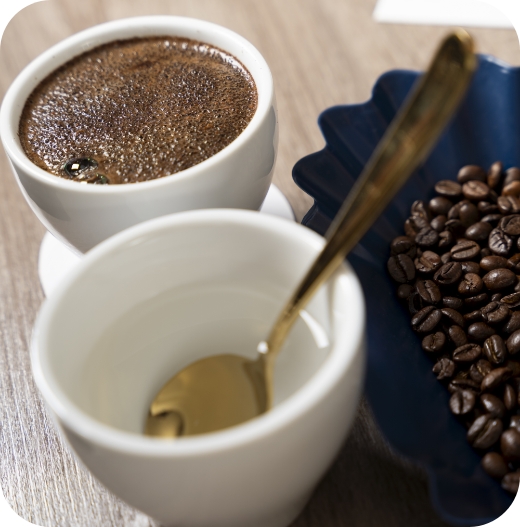 Nahaufnahme eines Tisches mit einer weißen Cupping-Schale voller gebrühtem Kaffee, einer leeren Schale mit einem goldenen Löffel und einer blauen, geriffelten Schale mit gerösteten Kaffeebohnen.