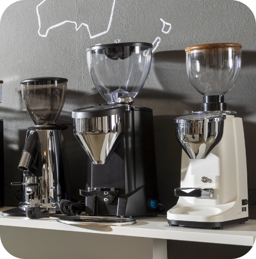 Drei professionelle, elektrische Kaffeemühlen (Schwarz, Chrom und Weiß) mit transparenten Bohnenbehältern stehen auf einem Regal vor einer dunkelgrauen Wand mit Weltkarten-Umriss.