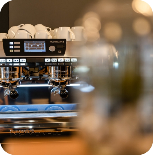 Nahaufnahme einer professionellen Siebträgermaschine (Nuova Simonelli Aurelia II) mit zwei Siebträgern und einem digitalen Display. Oben auf der Maschine stehen gestapelte, weiße Espressotassen.