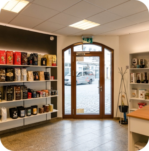 Eingangsbereich des JoKa Kaffee Ladengeschäfts mit einer braunen Glastür und beidseitigen weißen Regalen, die voll mit verpackten Kaffeebohnen und Zubehör sind.