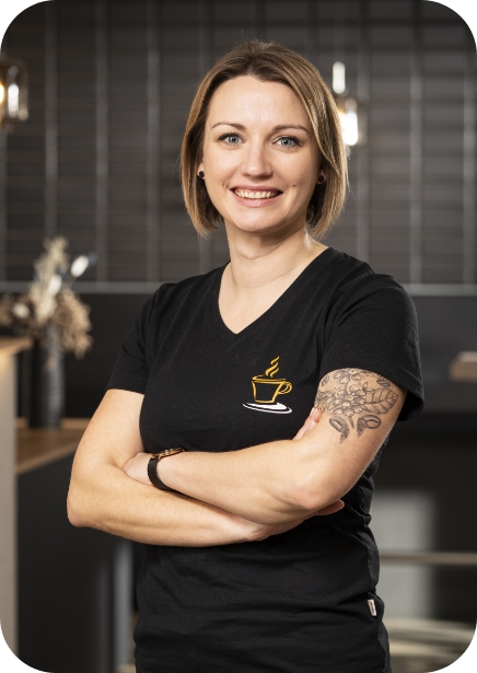 Professionelles Brustbild-Porträt von Ramona, einer lächelnden Mitarbeiterin von JoKa Kaffee, die ihre Arme verschränkt hält und ein schwarzes T-Shirt mit dem goldenen Logo sowie Tätowierungen am linken Arm trägt.