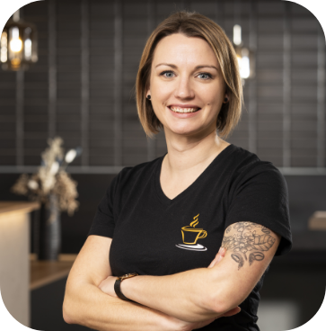 Professionelles Brustbild-Porträt von Ramona, einer Mitarbeiterin von JoKa Kaffee, die lächelt und die Arme verschränkt. Sie trägt ein schwarzes T-Shirt mit dem goldenen JoKa Kaffee Tassen-Logo und hat Tätowierungen am linken Arm.