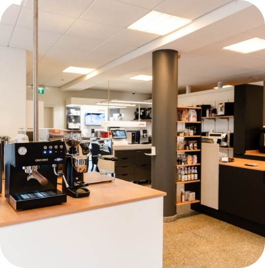 Innenansicht des JoKa Kaffee Showrooms mit einer schwarzen Espressomaschine (Ascaso) und einer Kaffeemühle im Vordergrund. Im Hintergrund sind Regale mit Kaffeeprodukten und weitere Kaffeemaschinen ausgestellt.