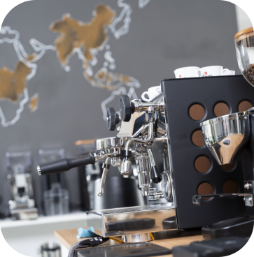 Professionelle, schwarze Siebträgermaschine und eine Kaffeemühle mit vollem Bohnenbehälter im Schulungsraum von JoKa Kaffee. Im Hintergrund ist eine Weltkarte an der Wand.