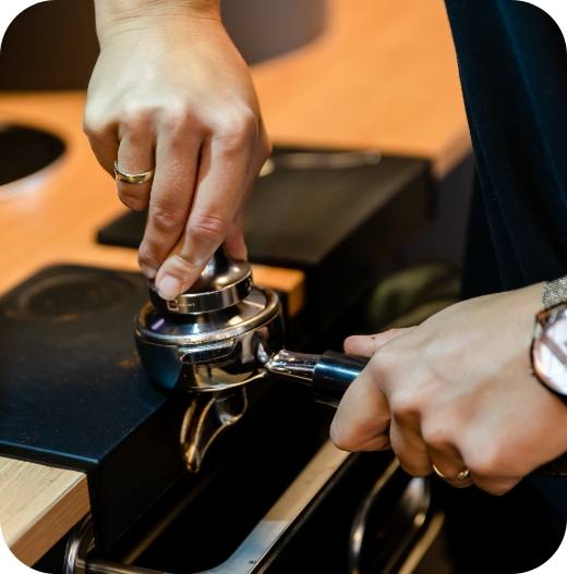 Nahaufnahme von Händen, die mit einem metallenen Tamper Kaffeemehl im Siebträger einer Espressomaschine auf einem schwarzen Tamping-Matte festdrücken.