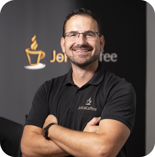 Professionelles Brustbild-Porträt von Torsten, einem lächelnden Mitarbeiter von JoKa Kaffee. Er trägt ein schwarzes Poloshirt mit dem JoKa Kaffee Logo und steht vor einem schwarzen Hintergrund mit großem Firmenlogo.
