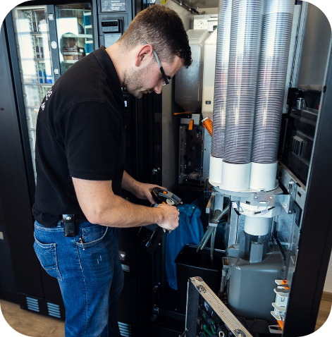 Ein Service-Techniker mit Brille, schwarzem Poloshirt und Jeans wartet oder befüllt einen Kaffee-Vending-Automaten. Die Becherstapel und der offene Innenraum des Automaten sind sichtbar.
