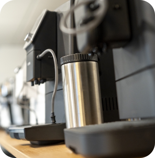 Nahaufnahme eines silbernen, isolierten Milchbehälters, der vor zwei schwarzen Kaffeevollautomaten (vermutlich Jura) steht. Die Geräte stehen in einer Reihe auf einer hellen Holztheke.